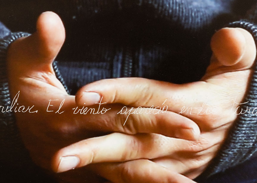 Detail of a photograph of hands with fingers interlaced with the palms open toward the body wearing a grey sweatshirt. There is cursive writing delicately etched into the middle of the photograph in a band from left to right; what we can see of the text in this detail reads, "...iliar. El viento aparecía en las..."
