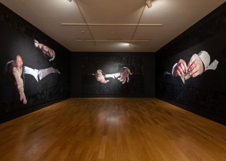 In the large gallery of Daniela Rivera's exhibition "Labored Landscapes (where hand meets ground)" we see three dark walls and a wood floor. Each wall has a single painting of a giant pair of hands; each pair of hands is gesticulating as in mid-conversation. The body to which the hand belong fades into the black canvas and the dark wall color.