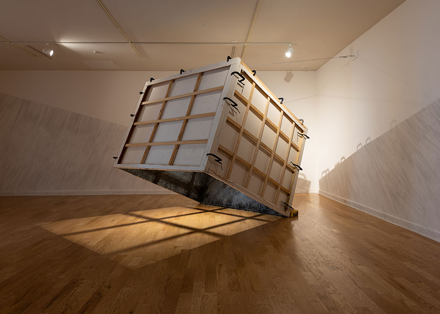 A cube made of stretched canvasses is titled at an angle so one of the canvasses is touching the wooden floor, and the others are suspended in the air. There is no top or bottom to the cube, and the light shines through the art onto the floor. There is a thick layer of ash on the inside of the canvasses, some of which has fallen onto the ground beneath the sculpture.