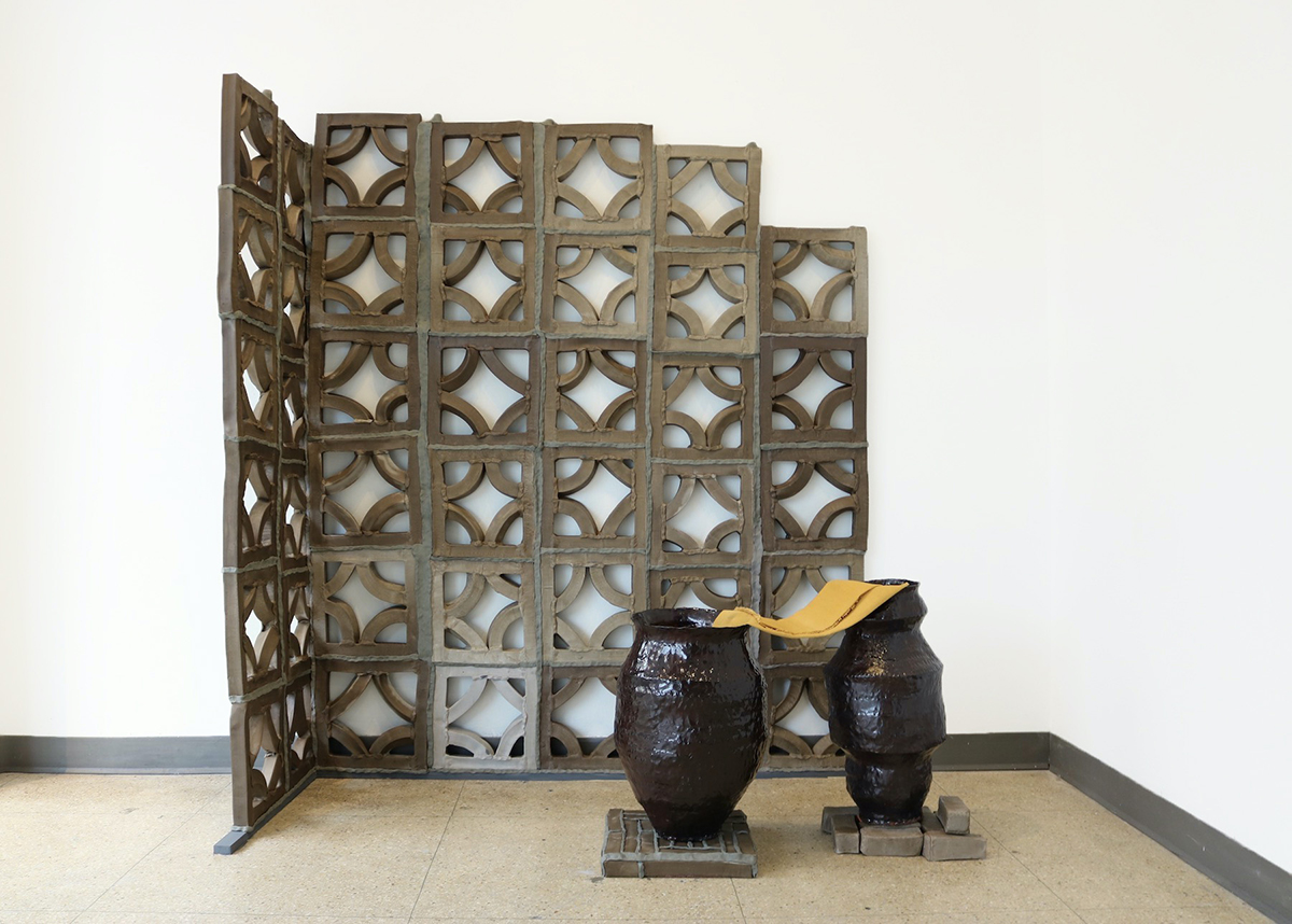 Two dark drown ceramic vessels rest in front of a tan-grey patterned ceramic wall. The vessels rest on low ceramic blocks and a yellow piece of linen is folded with each of its ends in each of the vessel openings. The ceramic wall creates a corner behind and to the side of the vessels; its rough-hewn lace-like sculpted pattern is made of squares, about a foot in each direction, with arcs in each of the corners that visually suggest circles with the other squares in the pattern.
