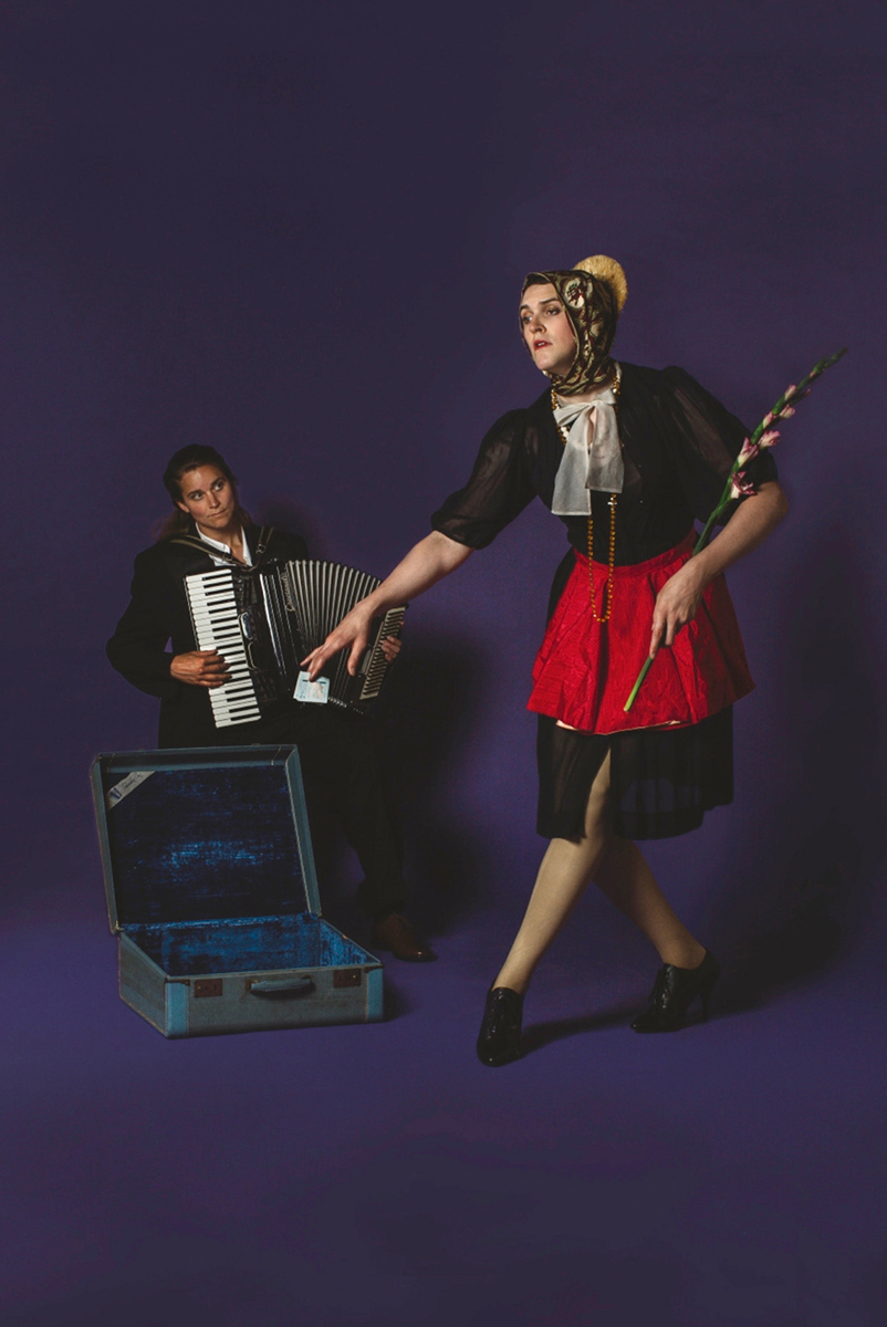 A tall fair-skinned white person poses gracefully as if dancing in a knee-length black dress, red apron, white neck bow, and floral head scarf with a stalk of flower in their left arm. To their left and behind in the image's frame, an accordion player, dressed in a black jacket and white shirt, sits in front of an open blue-velvet lined suitcase and watches the dancing person. The scene harkens to art historical images and is clearly set up in a studio in front of a vibrant deep purple backdrop.
