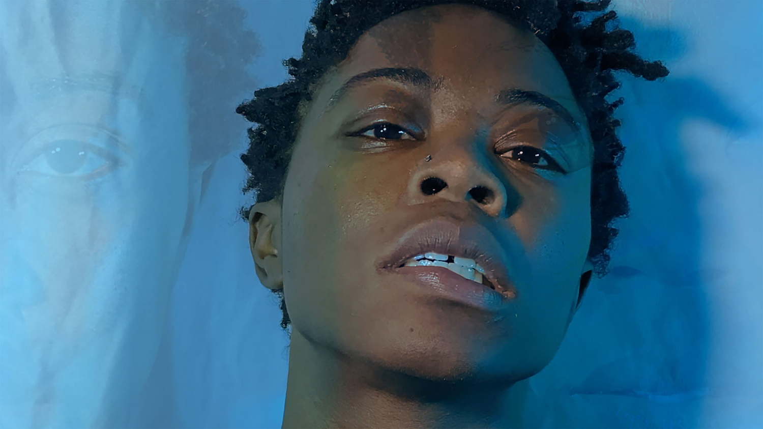 An African American woman looks directly into the camera — emotionally, and in the center of a cyan blue field — head tilted slightly back and to the side, with her mouth slightly open. Other, different, images of her face, are lightly overlaid to the left and to the right, giving the blue space of this film still a dynamic, watery feel of motion.