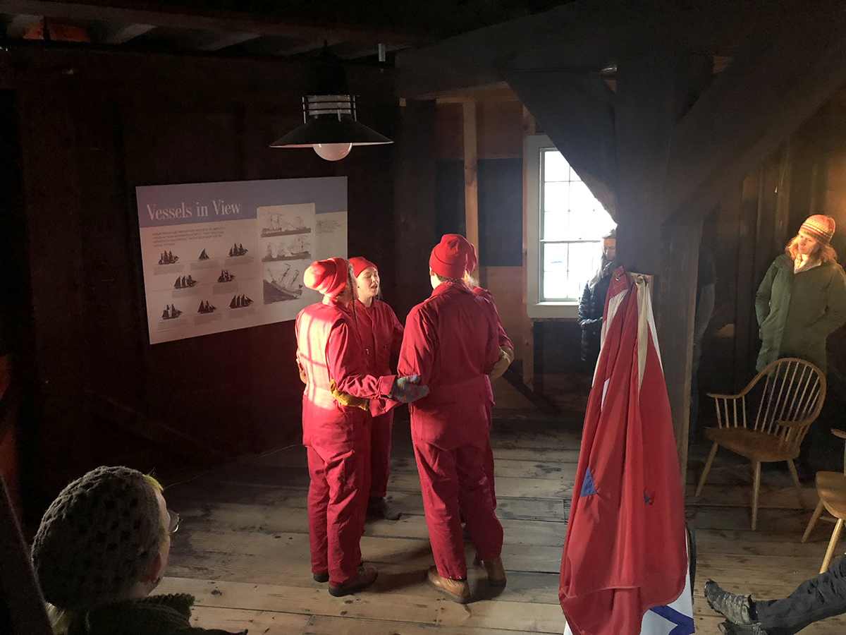 Four fair-skinned people in red utility jumpsuits and red knit hats hold each others' arms in a close circle in the center of a wooden-framed room. They stand next to an informational historical sign about vessels that one might be able to see from the observatory location. On the opposite side is a wooden beam, upon which several red and white cloths have been hung. Audience members can be seen at the periphery of this image, taken during the performance.