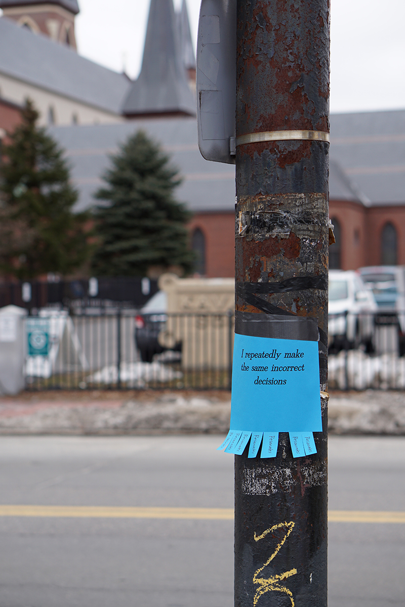 A lampost just off center of the frame has a blue paper sign taped to it, in the style of an advertisement with tear-off pieces at the bottom. The sign reads, "I repeatedly make the same incorrect decisions" in an elegant serif typeface. The tear-off pieces at the bottom each have the word "Resonates" handwritten on them in pen; two of the pieces have been taken.
