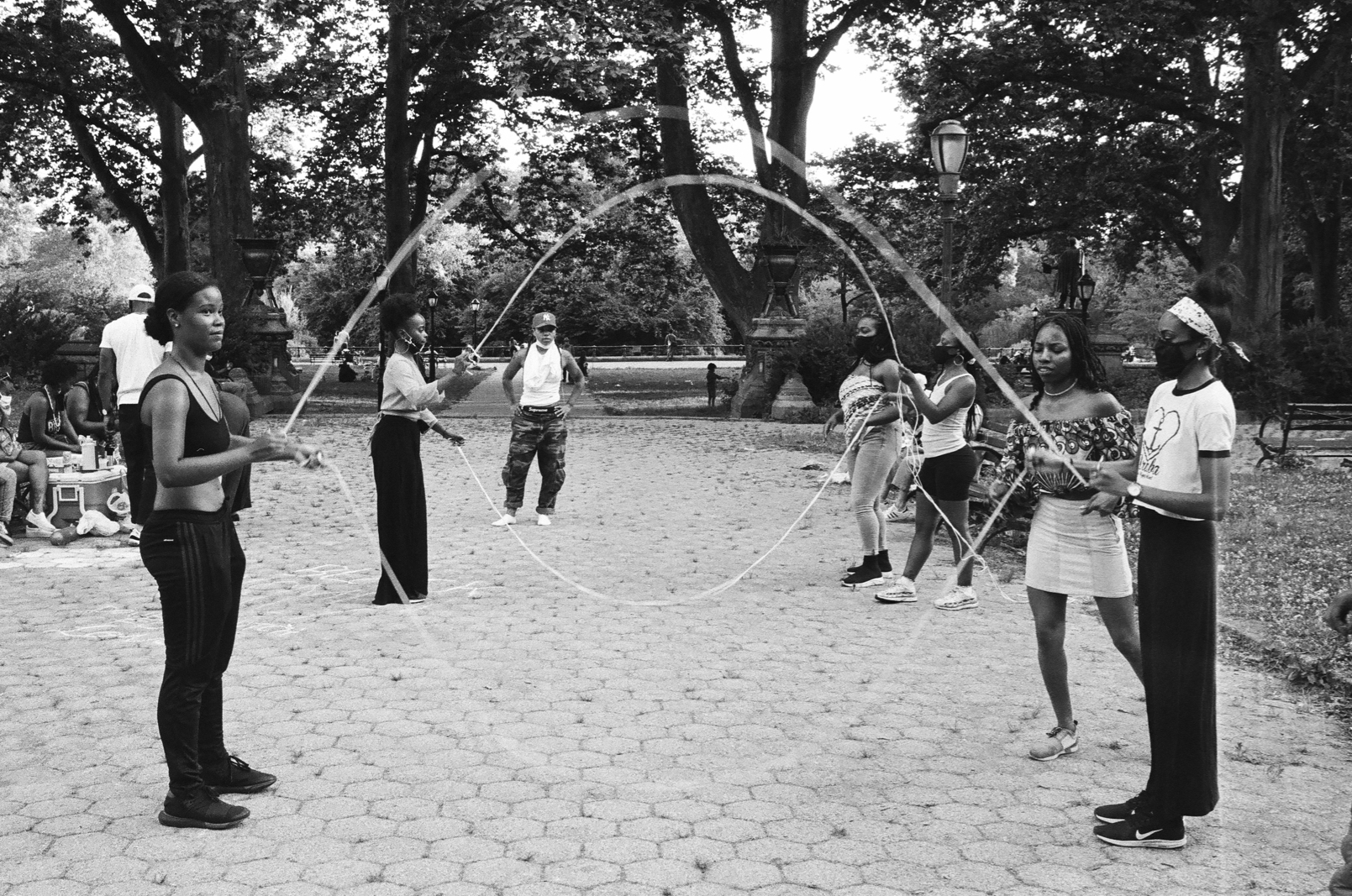 In this black and white photograph, the camera looks down the center of two sets of double-dutch jump rope, held by young Black women, some wearing face-masks in light of the coronavirus pandemic. Two of the women, standing on the right of the photograph, look ready to jump into each set of ropes.