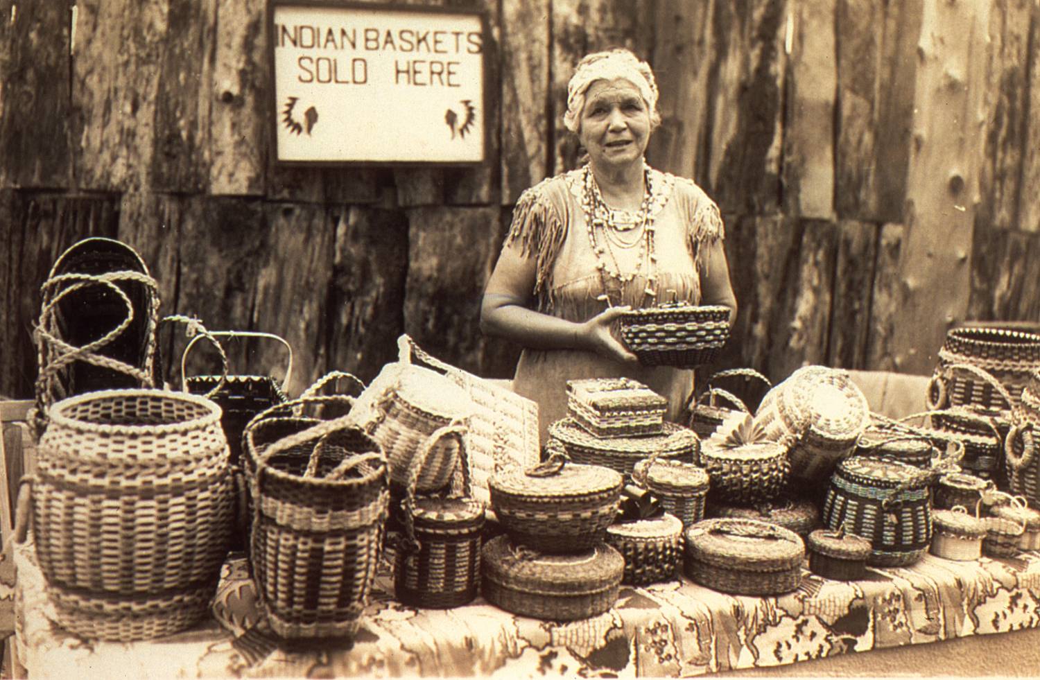 A Penobscot woman stands at a table with a large display of baskets spread out before her. She is holding one proudly in her hand. To the left of her, a sign on a wall of a building behind her says, "Indian Baskets Sold Here."