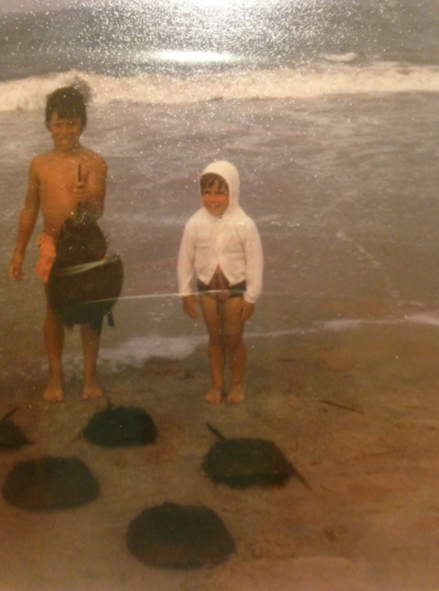 Two kids in a rephotographed image excitedly stand at the edge of the ocean's lapping waves with six horseshoe crabs. The taller child, on the left, proudly lifts one of the crabs by its telson. The younger child on the right, beaming in a white hoodie with hands pressed at their sides, poses happily behind five horseshoe crabs in a patterned formation, as if synchronized. 