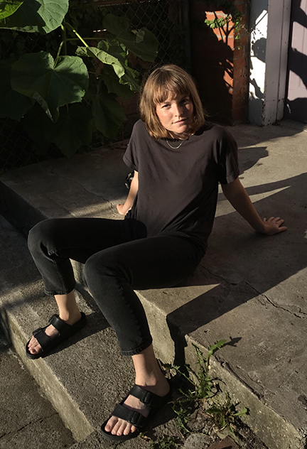 Erin Johnson is dressed in a black tshirt, black jeans, and black birkenstock sandals, sitting on a concrete step, leaning back on her hands. She's a fair-skinned white person with chin length reddish brown hair and is looking directly into the camera.