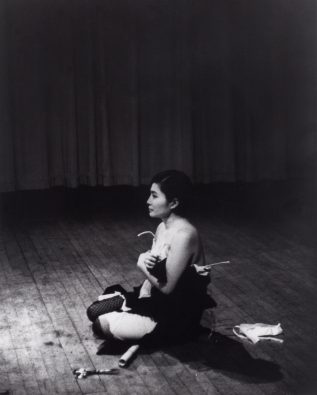 A black and white photograph of the artist Yoko Ono, who sits cross-legged on a wooden floor. She holds her dress to her body, which has been slowly cut away in small pieces by the audience.
