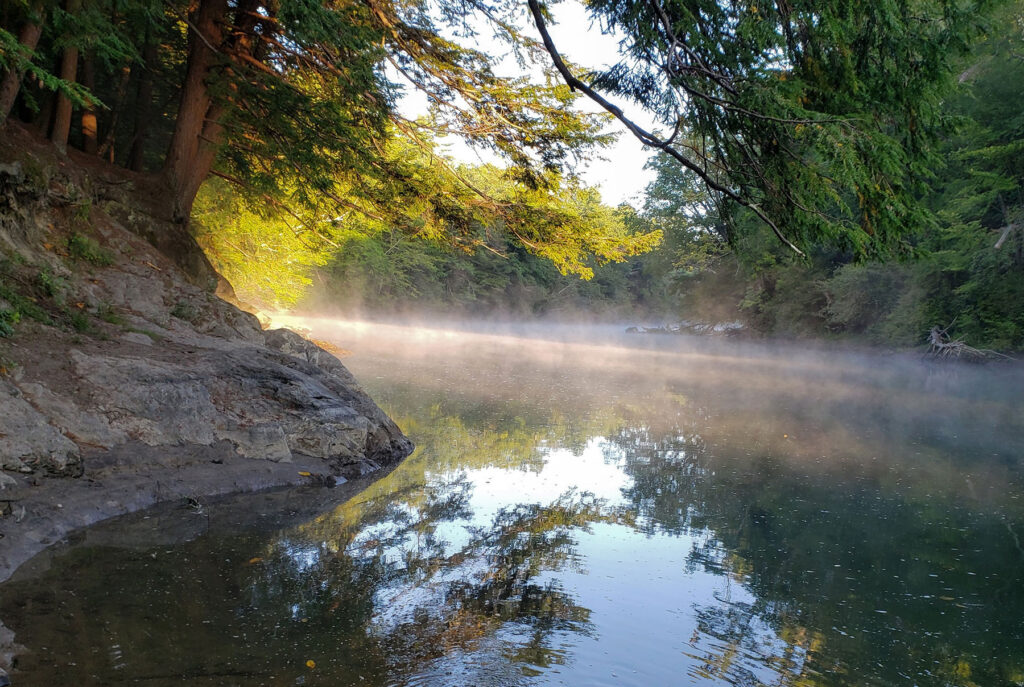 A view of a bend in the Presumpscot River just after dawn. Sunlight is just beginning to light up the trees that frame the rocky riverbanks, and there is a layer of white mist that sits on top of the dark water, which reflects the stones along the bank, the trees, and some sky above.
