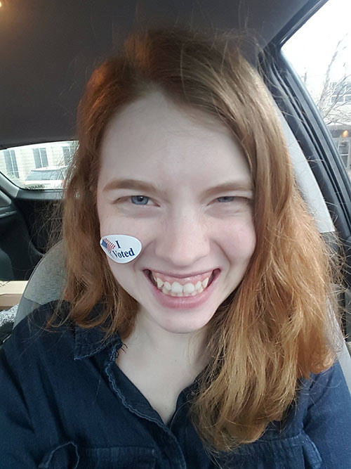 A pale-skinned white woman with blue eyes and shoulder-length red hair smiles into the camera. She is in a car, wearing a dark blue button-down shirt and sports an oval "I voted" sticker on her right cheek.