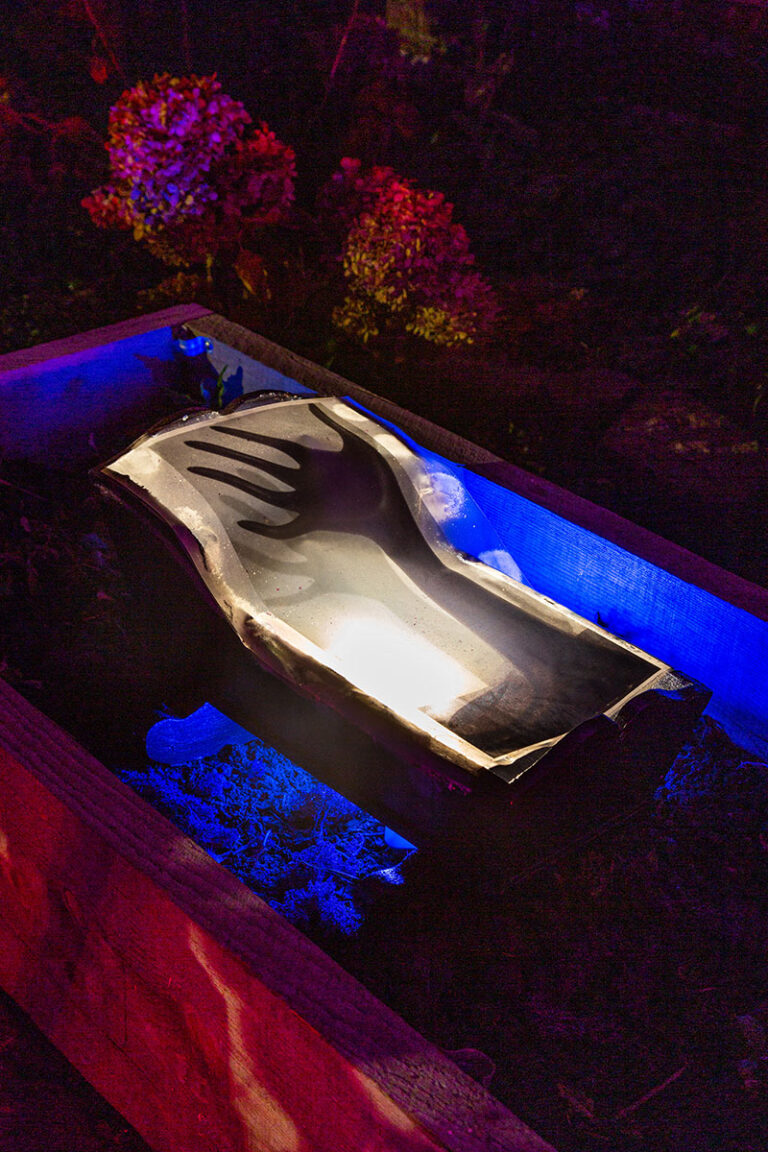 A white crib is nestled in a garden at night, with a black image of a reaching hand overlaid onto it. The crib is surrounded by otherworldly blue and pink light.