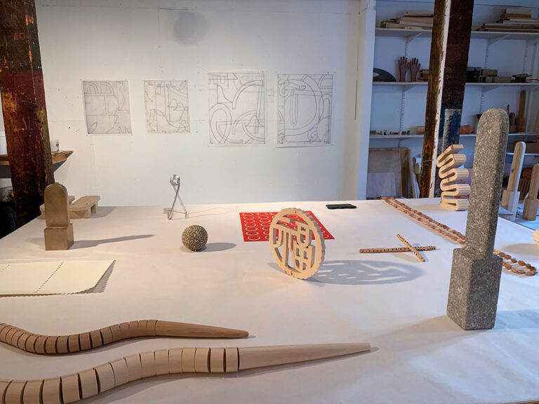 A glimpse of Elizabeth's Atterbury's studio, which features a white table with objects artfully arranged on it: segmented snakes, strings of wooden beads, an enlarged clothespin spring, wooden sandal soles, and more. On the wall behind the table are graphite drawings on white paper of some of these objects and other abstract shapes.